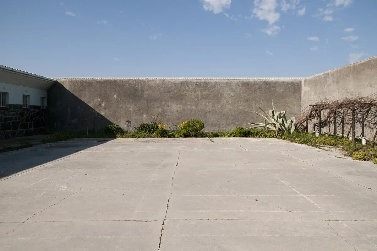 A walled concrete courtyard in Section B of the Maximum Security Prison on Robben Island, with plants growing along the base of the wall under a blue sky