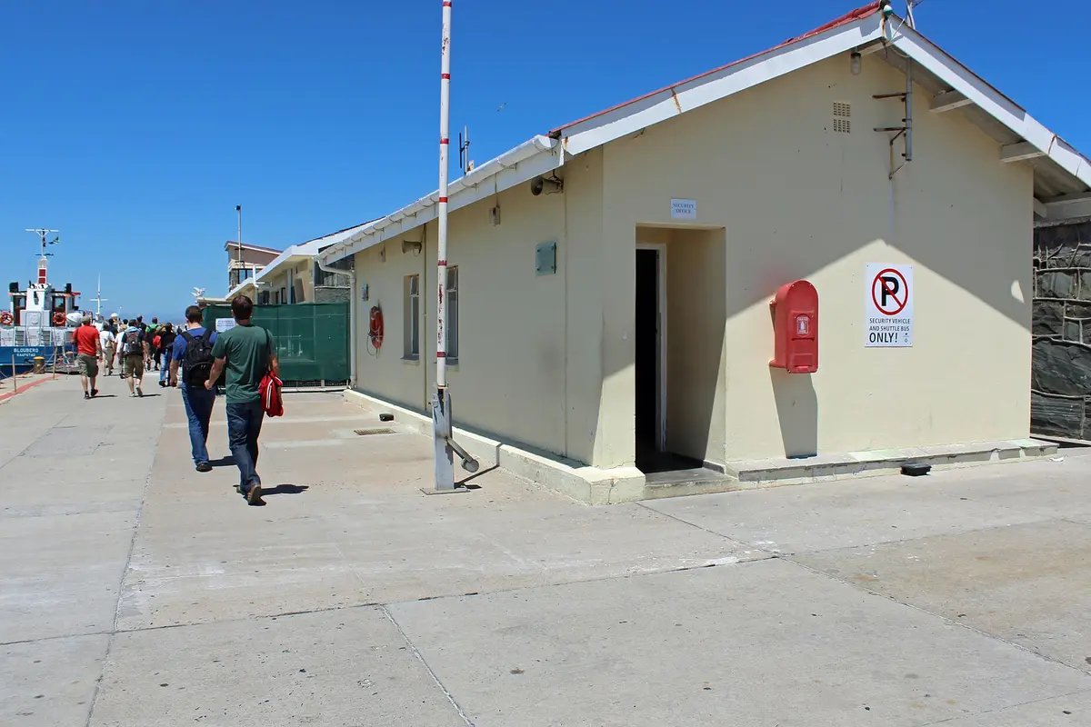 Cream-coloured harbour buildings on the quay at Murray's Bay Harbour on Robben Island, with visitors walking along the quay under a clear blue sky